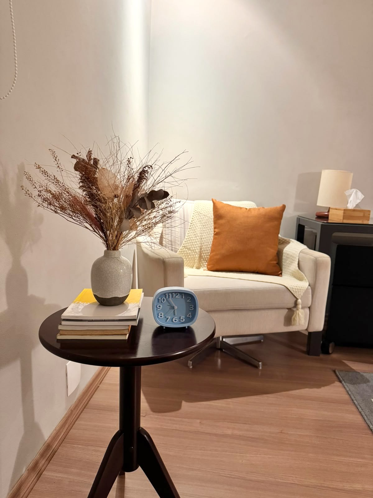 Detalhe do consultório: mesa lateral com flores secas, livros e relógio, e a poltrona ao fundo